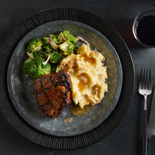 Dark black dinner plate with bacon mashed potatoes, broccoli and coffee rubbed grilled steak next to a black and silver fork on a dark background. 