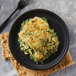 Creamed spinach with melted crisp cheese on top on a black plate on a brown napkin with a black fork on the side. 