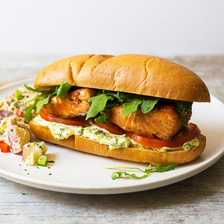 White dinner plate with a creole salmon sandwich with a fillet of salmon on a long roll with sliced tomatoes and creole sauce.