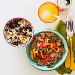 cuban style ropa vieja with rice and beans in colorful bowls on a white background with a can of orange soda in a glass