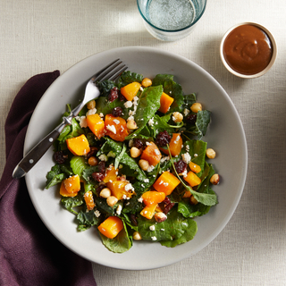 White dish filled with a green salad with chunks of butternut squash with a metal fork next to a glass of water and a dish of brown salad dressing. 