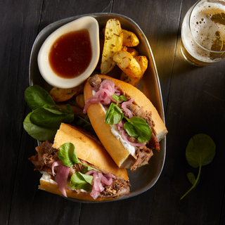 Square dish with a French dip sandwich sliced in half and jus in a pourable dish on the side with roasted potatoes and a glass of beer. 