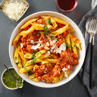 White bowl filled with garganelli pasta topped with sausage ragu, green dressing, shaved cheese with silverware on the side and a glass of red wine above. 