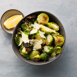 Garlic roasted brussels sprouts in a small dark bowl with shaved cheese on top next to a dish with a fresh lemon slice on the side. 