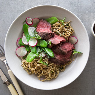 White dish with ginger garlic marinated flank steak sliced on top of wheat noodles with raw radish slaw on top, next to a fork and knife on a grey background. 