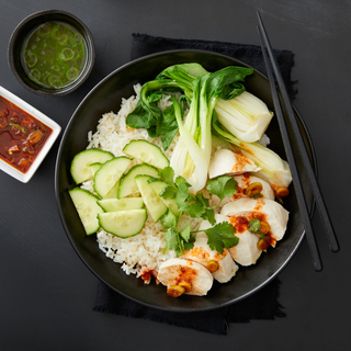 Ginger poached chicken breast on white rice with bok choy and sliced cucumbers in a black bowl with black chopsticks on a black napkin on a black table. 