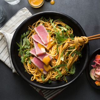 Overhead of a black dinner plate with grilled ahi tuna on a noodle salad with chopped greens and a small dish of pickles on the side. Chopsticks are swirled with some of the noodles in the dish. 