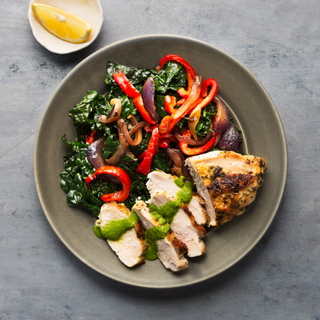 Sliced grilled chicken breast with roasted kale and bell peppers drizzled with green sauce on a black plate next to a white dish with a lemon slice. 