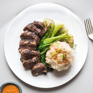 white round plate with sliced grilled red curry steak, sticky rice and grilled baby bok choy