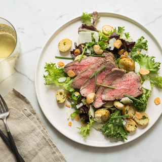 Grilled sliced tri-tip steak over a salad of potatoes and greens on a white dinner plate
