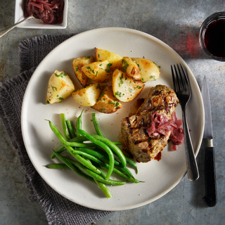 White round dinner plate with green beans, crispy potato wedges and a grilled tri-tip steak with a silver fork on a grey background. 