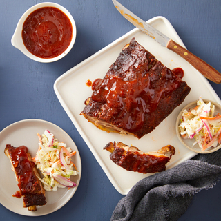 Square white plate on a blue background with a half rack of baby back ribs covered in barbecue sauce with a small plate holding one rib and coleslaw. 