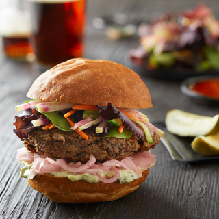 Side view of an impossible burger patty on a bun with pickled red onions, chopped vegetables and greens with a glass of soda in the background on a black wooden table. 