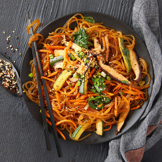 Korean japchae with sweet potato noodles, sliced cucumber and sesame seeds on top in a black bowl with black chopsticks and a grey napkin on the side. 