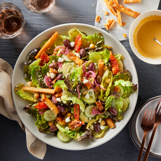 White dinner bowl filled with Jerusalem salad with chopped lettuce, sliced cucumbers, crispy naan strips, pickled red onions and dressing on the side in a small white bowl with two glasses of wine in the corner. 