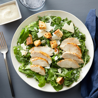 white bowl with shredded kale chicken caesar salad with sliced chicken breast, cheese and croutons