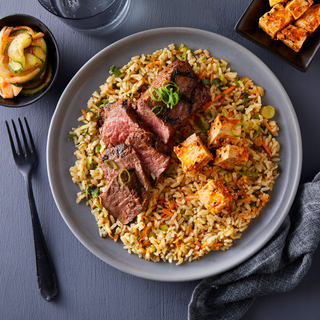 Blue dinner plate filled with brown rice salad topped with sliced Korean BBQ bavette steak with a black fork and a bowl of pickles on the side. 