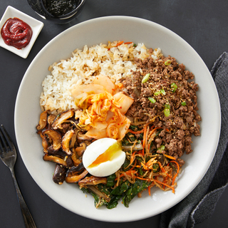 Korean bibimbap beef bowl with white rice, ground beef and a poached egg and vegetables on a white plate with a side of red-hot sauce in a small square dish. 