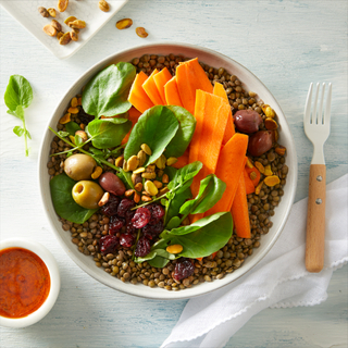 White bowl of lentils, sliced carrots, olives, baby spinach, nuts and seeds on a light blue background with a white napkin on the side. 