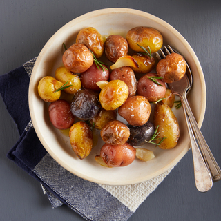 White bowl of roasted whole potatoes with fresh rosemary and a fork and spoon on the side. 