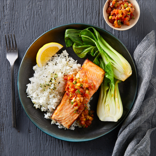 Black ceramic bowl with white rice, cooked bok choy halves and a salmon fillet topped with lomi-lomi tomatoes on a black wooden table. 