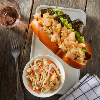 Rectangular white dinner plate with a New England style shrimp roll on a hot dog bun with a bowl of crunchy slaw on a wooden table top with a glass of rose and a checkered napkin. 