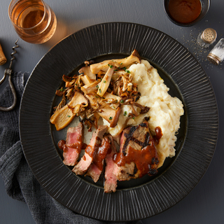 Black round dinner plate with New York steak sliced into thin pieces with mushrooms and mashed potatoes next to a glass of rose wine with a black napkin. 