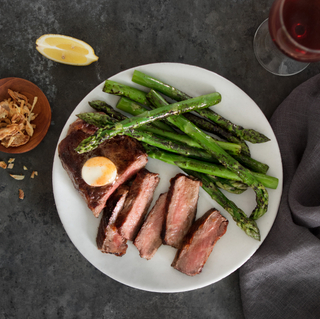 White bowl of New York strip steak sliced into five pieces with green asparagus on a dark background with a glass of red wine and a lime wedge on the wide. 