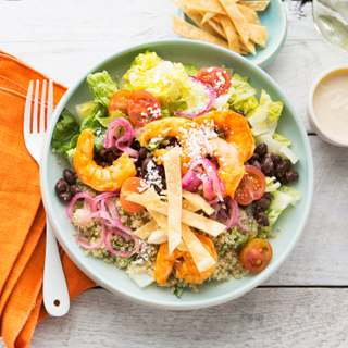 Green bowl filled with shrimp taco salad with colorful vegetables, crispy tortilla strips and roasted shrimp next to a folded orange napkin with a fork on a white table. 