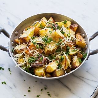 Parmesan roasted potatoes in a metal dish with handles topped with cheese and green herbs on a white marble table. 