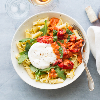 White bowl of pasta with tomato sauce and a ball of burrata with greens, a fork next to two glasses and a bottle of rose wine and a white napkin on a blue background. 