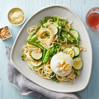 Pasta primavera with spaghetti noodles, a ball of burrata and fresh green spring vegetables on a white plate with a grey linen napkin on the side on a white wooden table. 