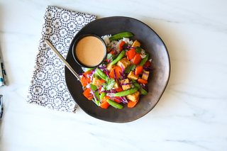 Black plate with peanut tofu stir-fry with red bell peppers, tofu and green beans with peanut dressing on the side next to a blue patterned napkin. 