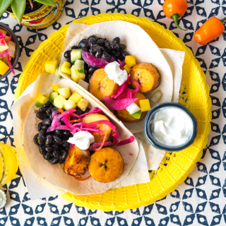 Bright yellow late with two tortillas holding sliced plantains, black beans, pickled red onions and diced mango salsa with a side of sour cream on a blue patterned background with three orange peppers on the side. 