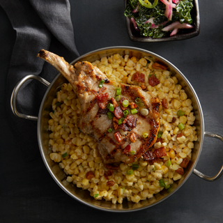 A silver dish with handles containing creamed corn and a large grilled pork chop with spicy pecans on top served with a square plate of kale salad on the side. 