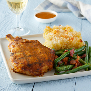 Square white plate with spiced grilled pork chop next to a square of mac and cheese with a crispy top and green beans with bacon in front of a glass of white wine on a white wooden table top. 