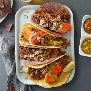 Rectangular plate with four red chili beef short rib tacos in two corn tortillas topped with carrots, limes and green and red salsa on a blue background with tortilla chips on the side. 