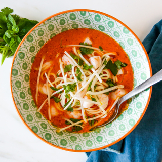 Red curry shrimp soup topped with matchstick vegetables and fresh herbs in a green and white patterned bowl with a fork in the bowl. 