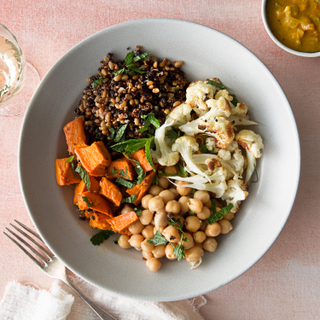 White dinner plate with red quinoa, dill chickpeas, roasted cauliflower and roasted sweet potatoes on a pink background with a silver fork and white napkin