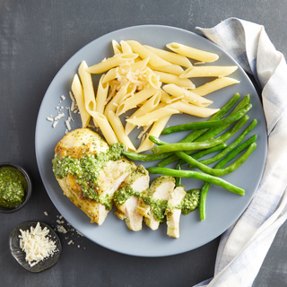 Roast chicken breast sliced with a drizzle of pesto sauce on top next to plain penne pasta and steamed green beans on a blue plate next to a white cloth napkin. 