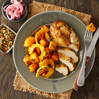 Roast chicken breast sliced on a plate next to roast winter squashed with a small dish of pickled red onions on the side and fork and knife on the plate. 