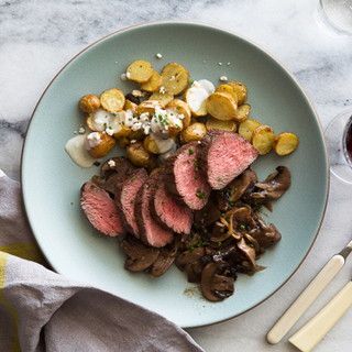 Teal dinner plate with balsamic onions, sliced bistro fillet steak and crispy potatoes with white dressing on a marble background. 