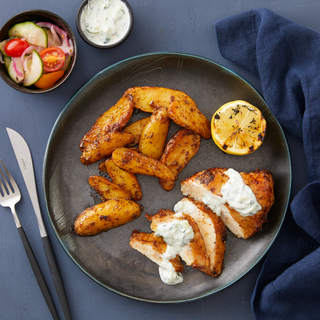 Roasted Greek chicken breast sliced into smaller pieces on a round black dinner plate with crispy potatoes and a small salad in a dish on the side on a dark blue background. 