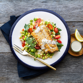 white plate with grilled cod with corn succotash on a wooden background with lemon and sauce on the side