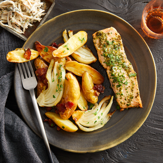 Brown dinner plate with roasted salmon fillet with green sauce next to crispy potato wedges and roasted sliced fennel with a fork. 