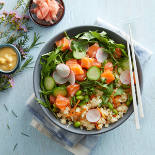 Blue bowl of salmon poke rice bowl with white chopsticks, chopped fresh salmon, radishes and greens on a light blue background with a folded napkin. 