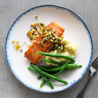 White dinner plate with blue edges holding green beans, roasted salmon, mashed potatoes and caper relish on top with a fork on the side on a grey background. 