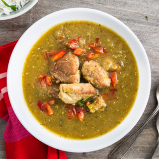 Santa fe-style green chile stew with pieces of chicken and chopped red peppers in a white bowl with a red napkin on a wooden table. 