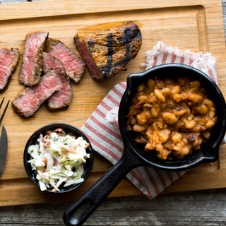 Wooden rectangular cutting board with Santa Maria BBQ tri tip steak sliced into five pieces with a mini cast iron skillet holding bacon ranchero beans and a small dish of coleslaw. 