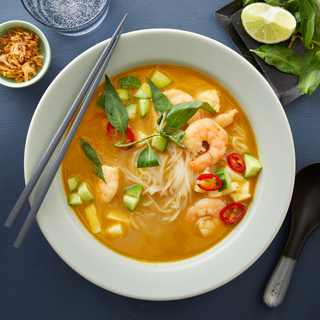 White soup bowl with shrimp laksa with chopped cucumber and fresh herbs on top with black chopsticks and fresh lime on the side. 